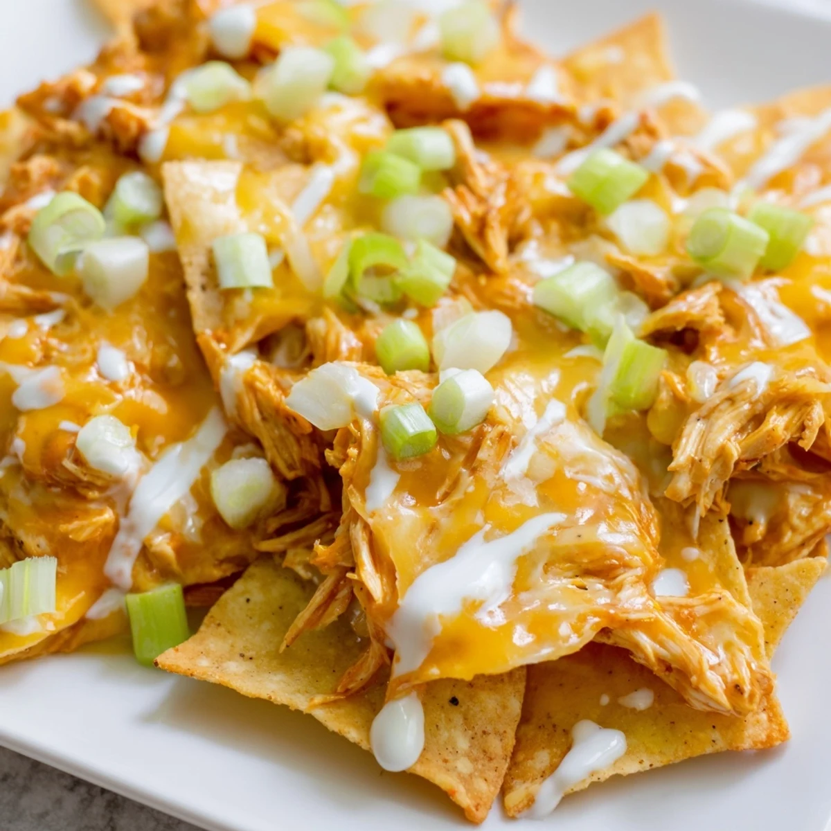 A close-up of Buffalo Chicken Nachos with golden melted cheddar and Monterey Jack over spicy shredded chicken, drizzled with creamy ranch and topped with fresh green onions and celery.  