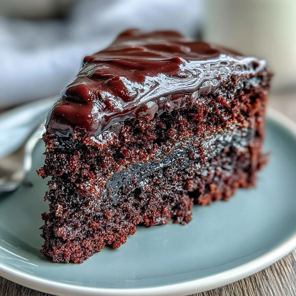 Easy Chocolate Fudge Cake with glossy icing sits beside fresh raspberries and a steaming cup of coffee on a table.