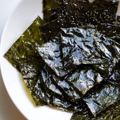 Golden-brown roasted seaweed chips stacked neatly on a bamboo tray, showcasing their light, crispy layers and scattered sesame salt crystals for a simple, savory vegan snack.