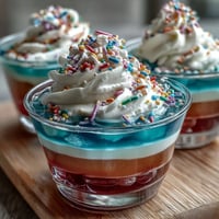 Rainbow Jello Cups for Kids: colorful layered dessert in clear cups with whipped cream and sprinkles, perfect for festive parties.